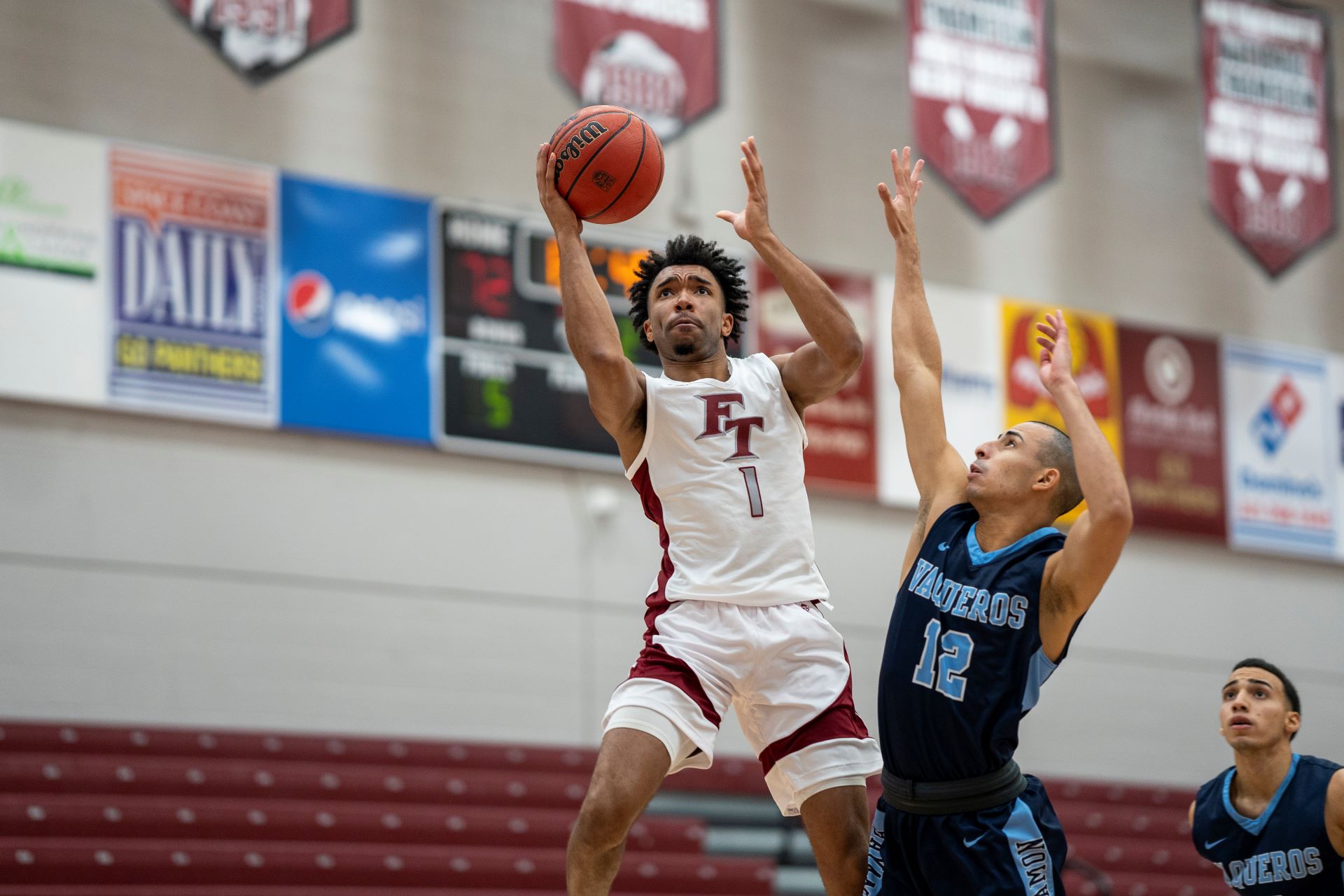 A Florida Tech basketball player in a white jersey with the number 1 going for a layup during a game against a player in a blue jersey with the number 12. Banners and advertisements are visible in the background